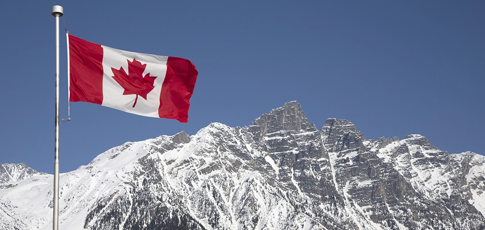 Canadian flag flying in front of snow-covered mountains under a clear sky