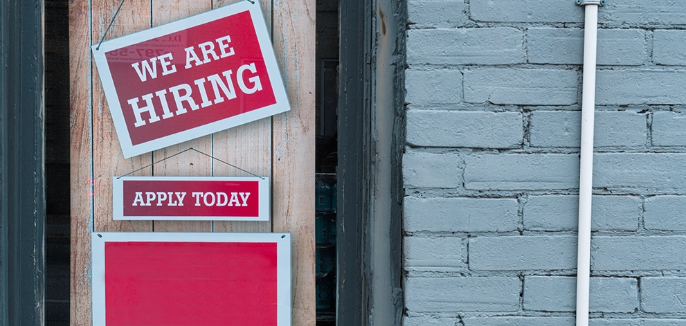We are hiring sign displayed in a shop window with apply today message