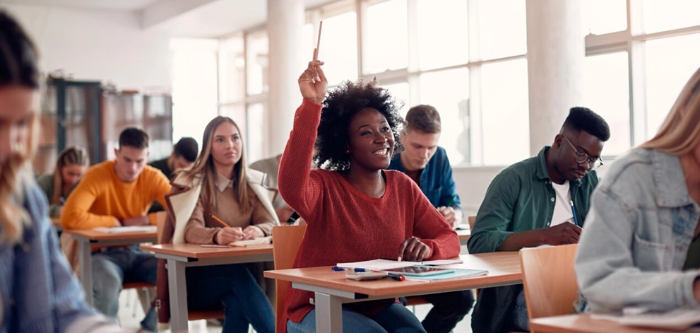 Student raising hand in a classroom while others take notes during a lesson