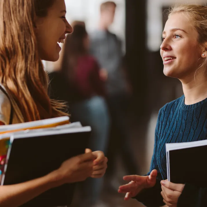 Two female students talking in a hallway, holding books and notebooks