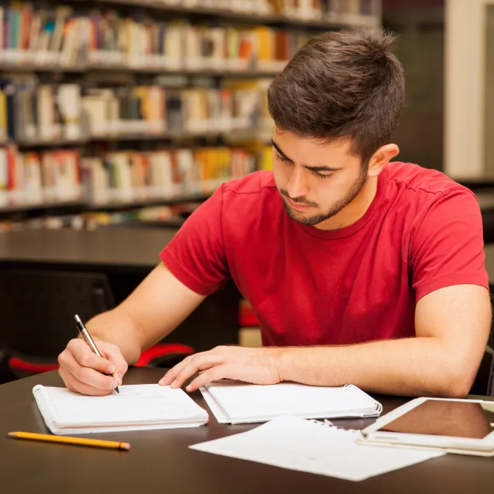 Man studying in a library with notebooks and an electronic device on the table