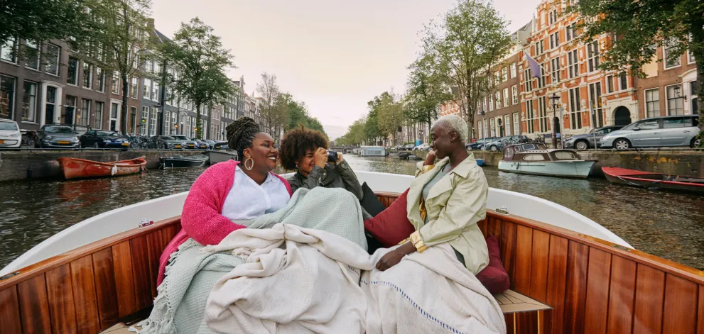 Friends sitting on a wooden boat wrapped in blankets during a canal trip