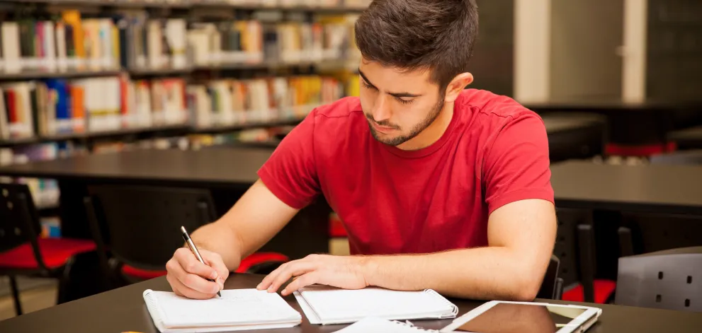 Man studying in a library with notebooks and an electronic device on the table