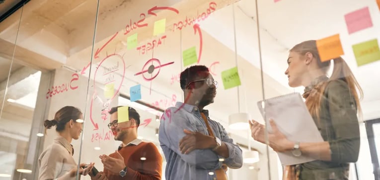 Group of people in a stand-up meeting discussing notes on a glass wall