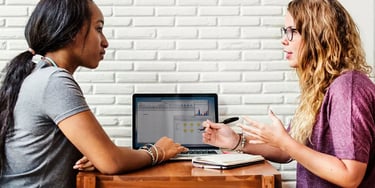 Two female students discussing charts on a laptop at a wooden table